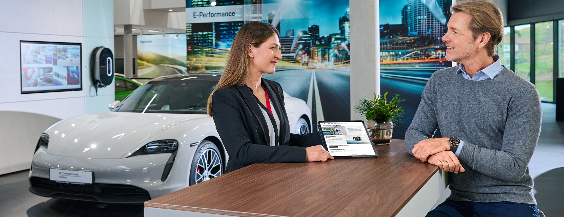 Ein Kunde sitzt an einer Theke im Porsche Zentrum und spricht mit einer Porsche Mitarbeiterin.
